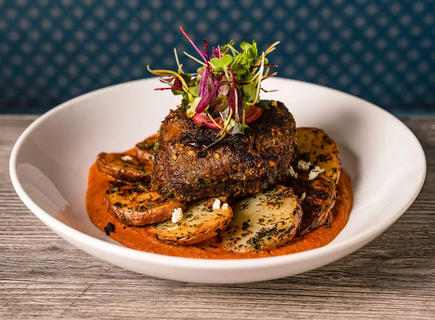 A white ceramic serving bowl displays a filet mignon sitting atop round potatoes and an orange vindaloo sauce on top of a wooden tabletop at CalilChili, one of the best Indian restaurants in Los Angeles, on OpenTable.