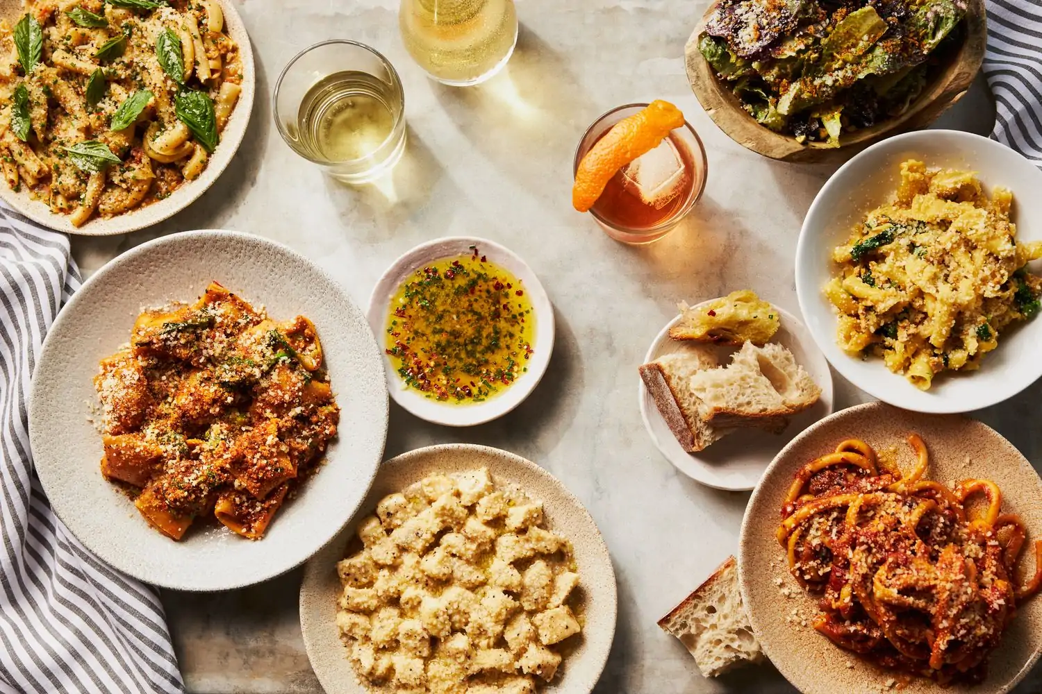 Five plates holding generous servings of pasta, including gnocchi with a cream-based sauce and spaghetti with a ragu, a salad, a plate of sliced bread, and a dish of oil at MIDA in Boston. Book on OpenTable.