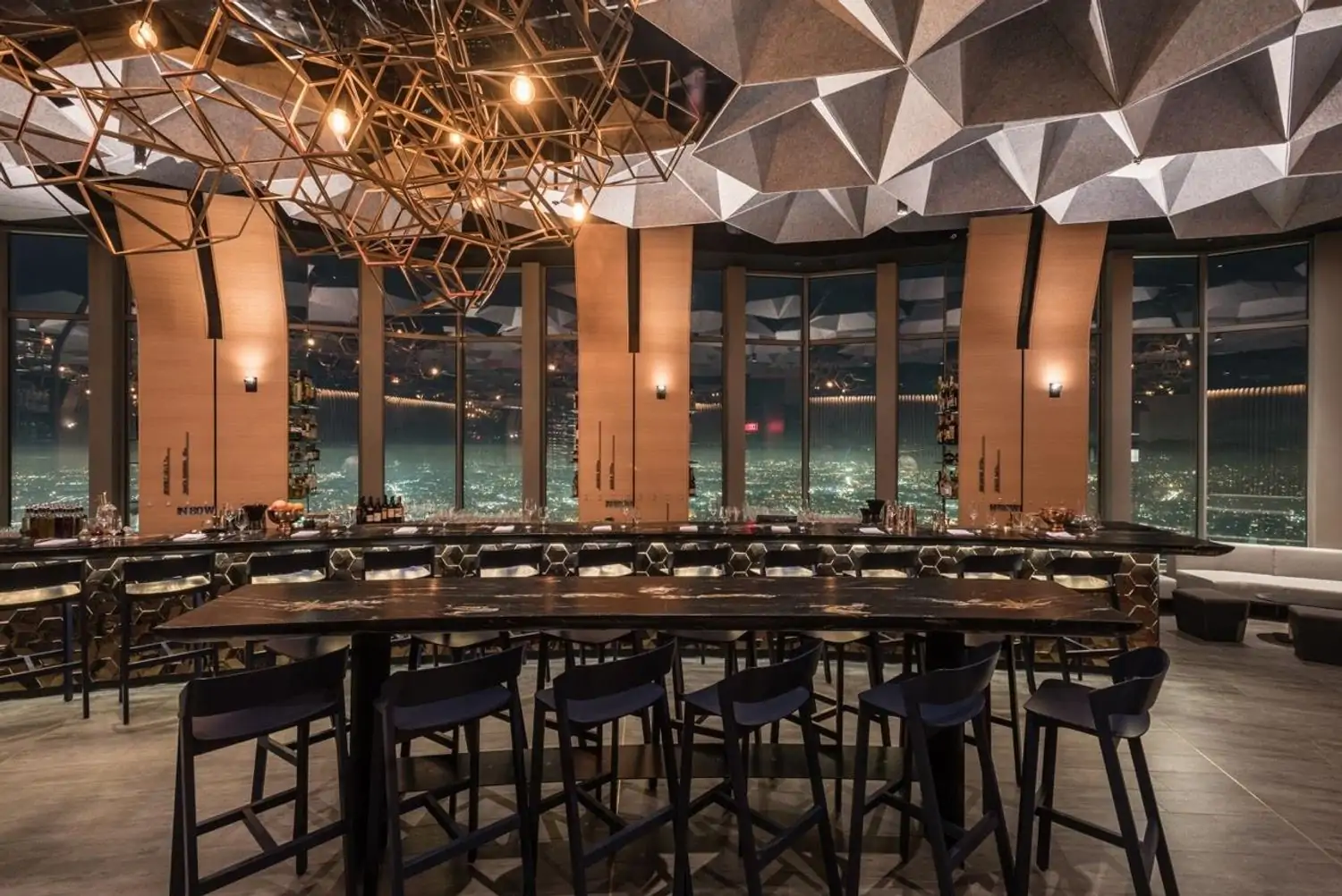 A dining room accented by cool shades of gray with high-top tables and chairs looking out over the city of Los Angeles at night at 71Above, available via OpenTable.