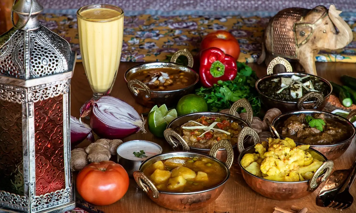 Six silver traditional Indian bowls with handles filled with food like curry, saag paneer, and cauliflower and a tall glass of mango lassi surrounded by fresh produce like red onion, ginger, red bell pepper, sliced limes and cilantro on a wooden tabletop at All India Cafe. Book on OpenTable.