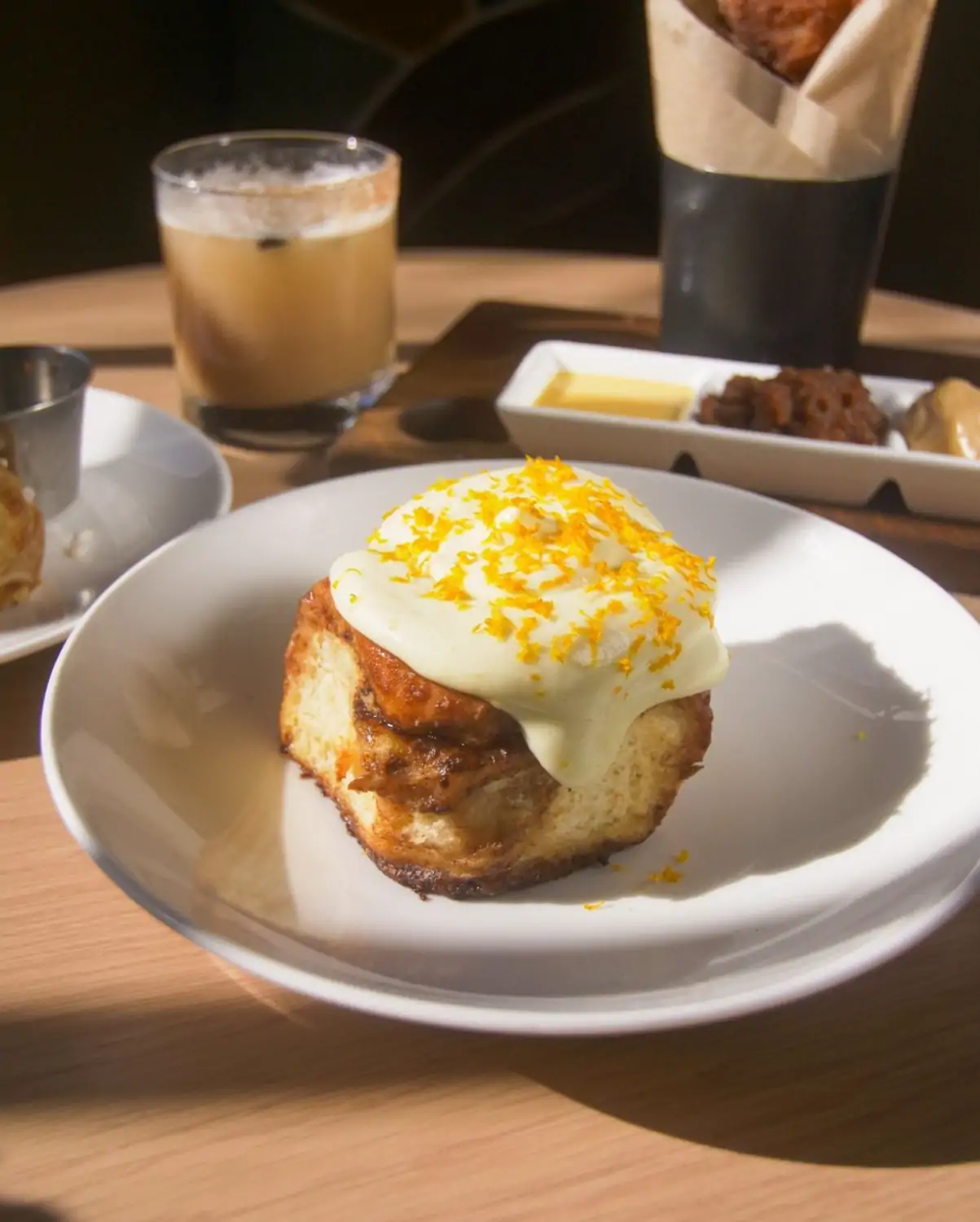 A plate containing a fresh cinnamon roll topped with frosting and garnish surrounded by an assortment of sauces and an iced coffee in the background at Manhattan Beach Post, one of the best American restaurants in Los Angeles, bookable on OpenTable.