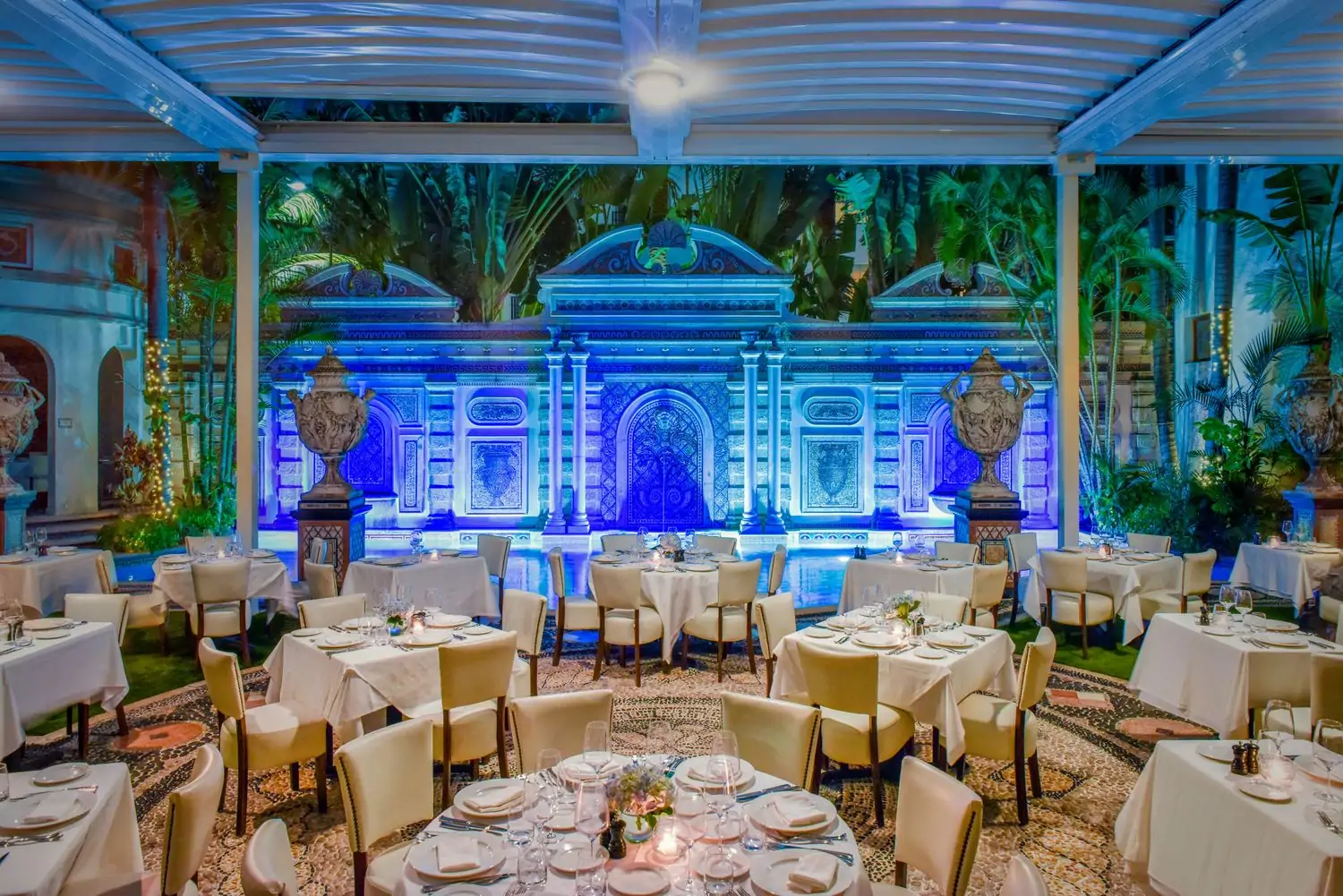 An al fresco dining area with a circular mosaic on the floor, 14 tables with pristine, white tablecloths, and a pool lit up in blue at Gianni’s at the Former Versace Mansion, one of the best restaurants in Miami Beach, on OpenTable.