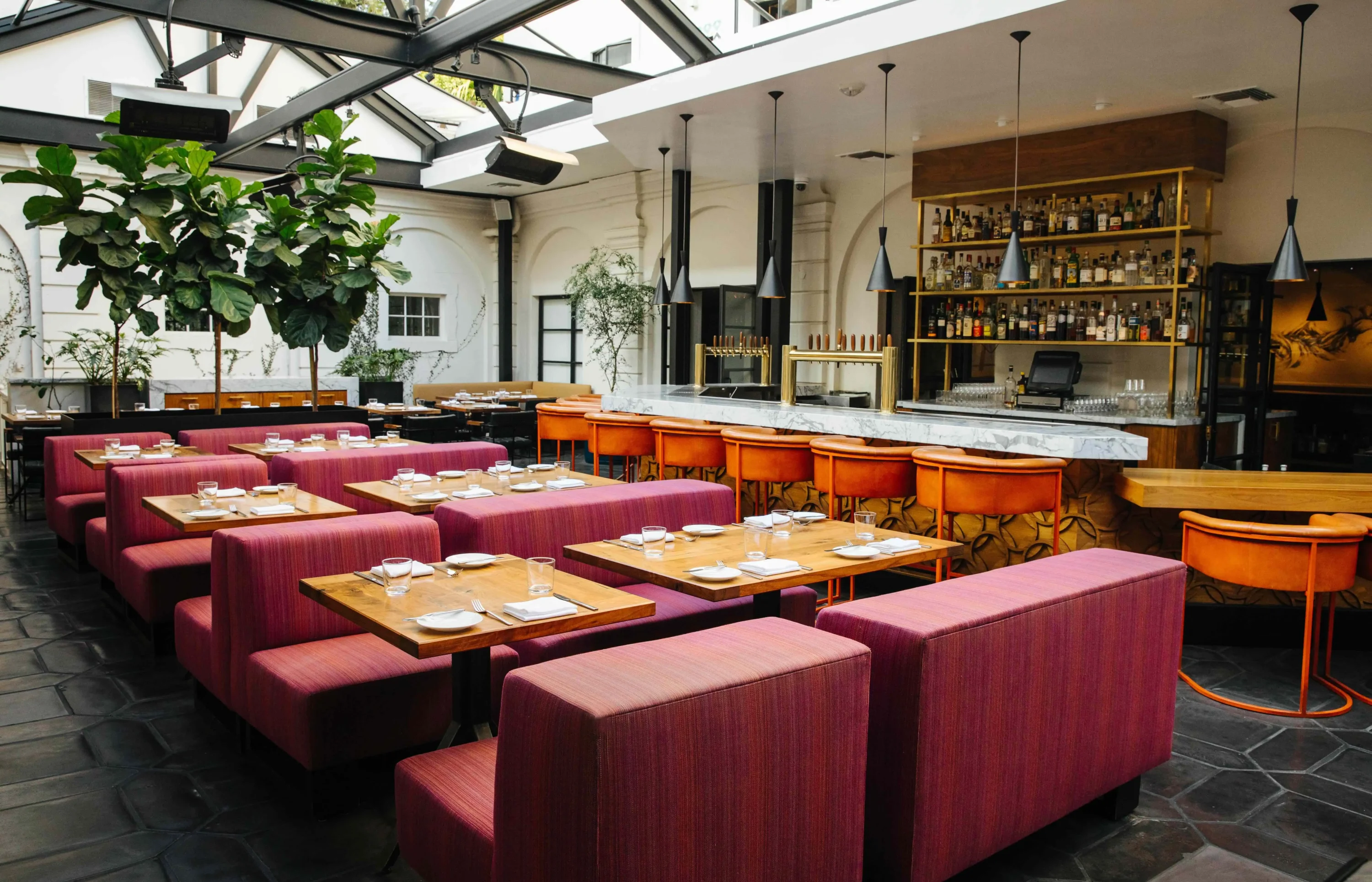 A dining room with a glass ceiling letting in plenty of sunlight with a full bar and red booth seating at Redbird, one of the best American restaurants in LA you can find on OpenTable.
