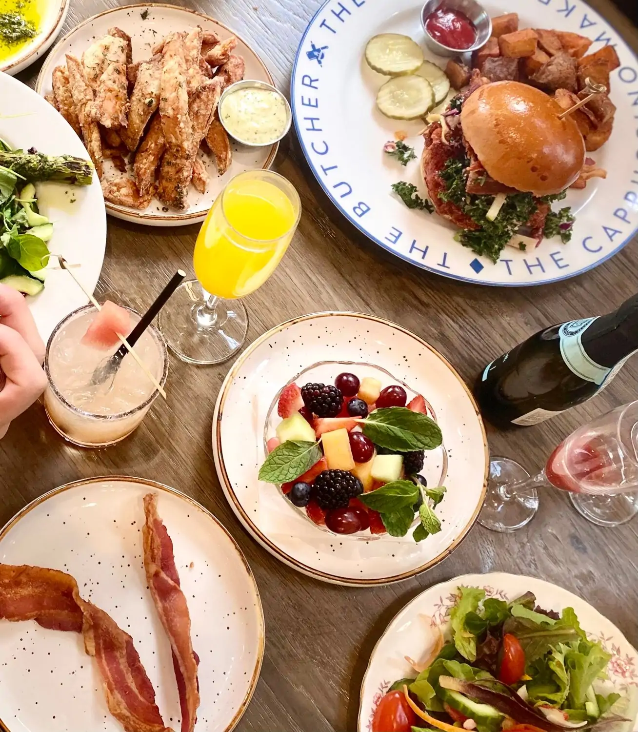A table setting with plates of fresh fruit, bacon, salad, fries, and a burger surrounded by colorful cocktails at The Butcher, The Baker, The Cappuccino Maker, a superb American restaurant in Los Angeles, CA, on OpenTable.
