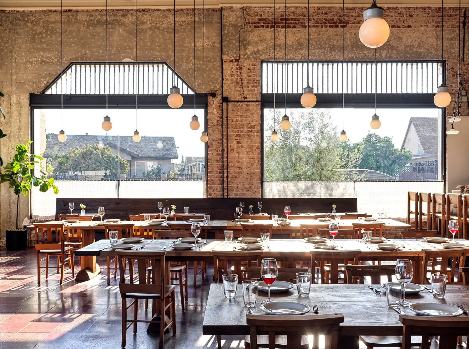 A dining room featuring exposed brick walls and minimalistic table settings in front of a large window through which a generous amount of sunlight lights up the room at Dunsmoor, bookable via OpenTable.