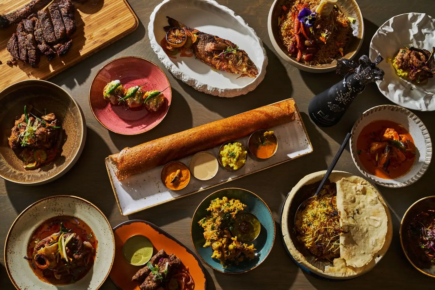 A wide variety of minimalistic plated Pakistani dishes, including whole fish, biryani, chaat, in contemporary stoneware, and sliced steak on a cutting board on top of a clean, modern wood tabletop at Shor. Book on OpenTable.