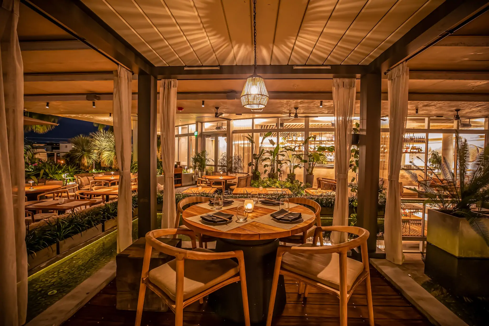 The rooftop terrace and open-air dining space with wooden tables and lush plants at MILA in Miami Beach. Book on OpenTable.