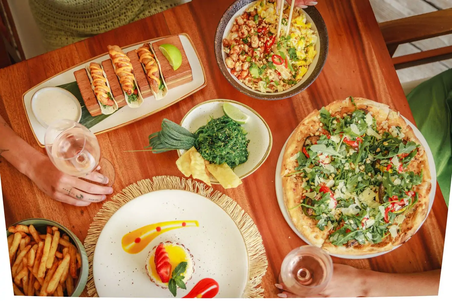 A wooden table with five plates of food, including pizza, a seaweed salad, French fries, and fried fish tacos, at Nikki Beach in Miami Beach, a restaurant available on OpenTable.