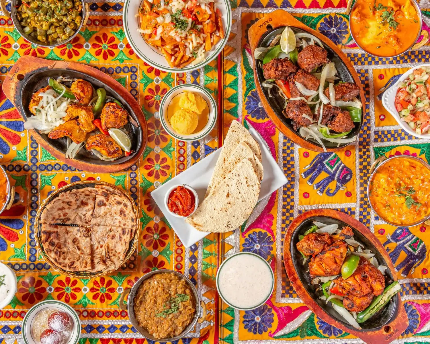 A colorful tabletop displays 14 Indian dishes, including raita, naan, tandoori chicken, gulab jamun, and more, at Electric Karma, one of the best Indian restaurants in Los Angeles at OpenTable.