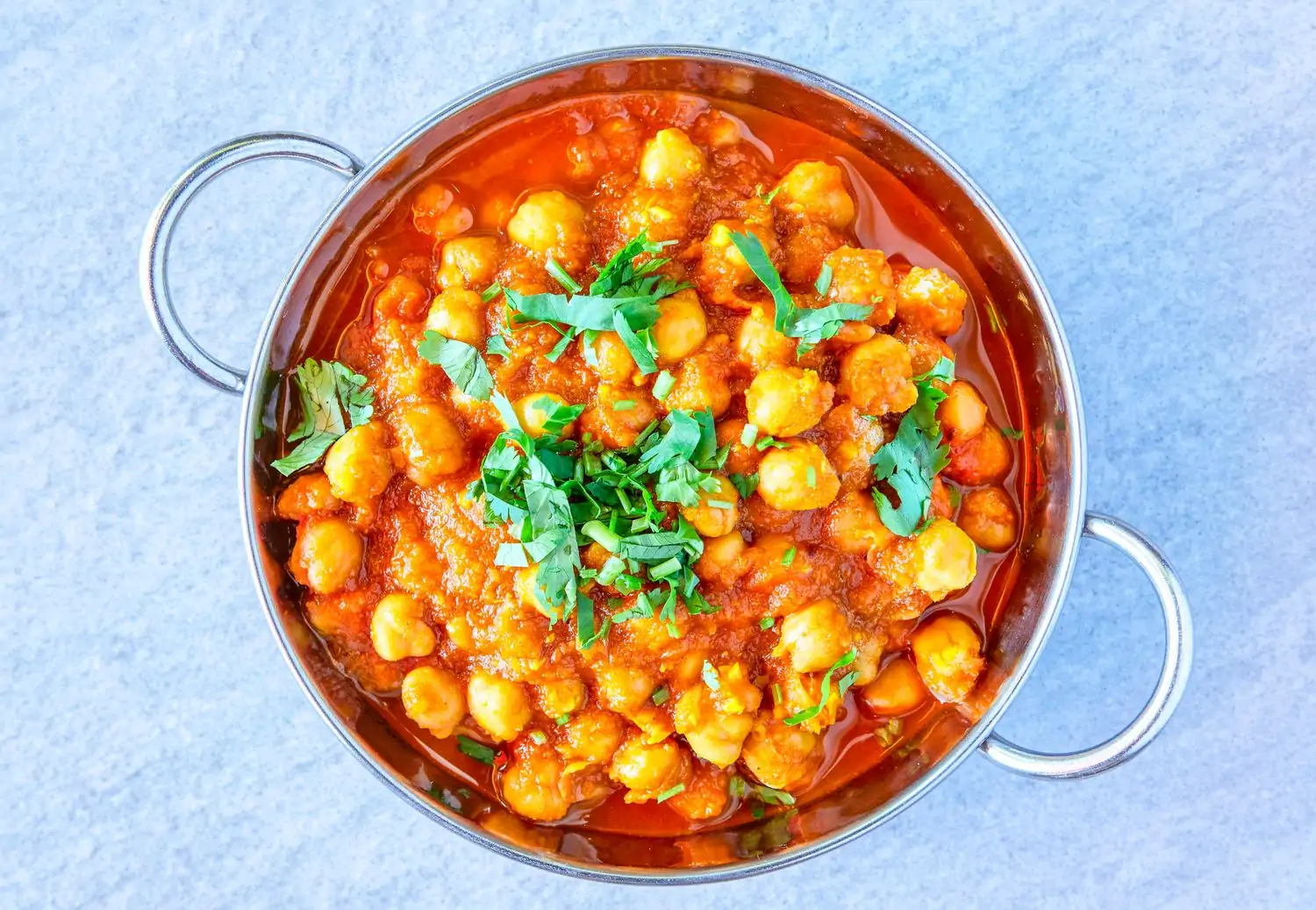 A silver bowl with handles filled with chickpeas in a red sauce topped with cilantro garnish rests on top of a blue marbled tabletop at Roots Indian Bistro in West Hollywood. Book on OpenTable.