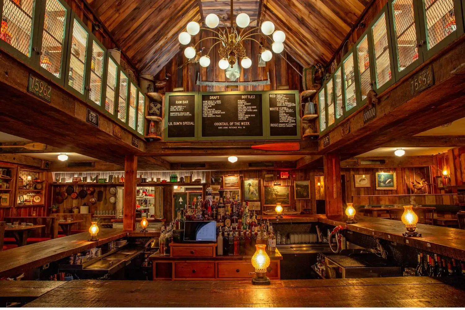 The hunting lodge-inspired interior at Hunter’s, one of the best warm and cozy restaurants in Boston, with reclaimed wood panels, a huge central bar, warm lighting from a chandelier, and oil painting depicting hunting scenes – book via OpenTable.
