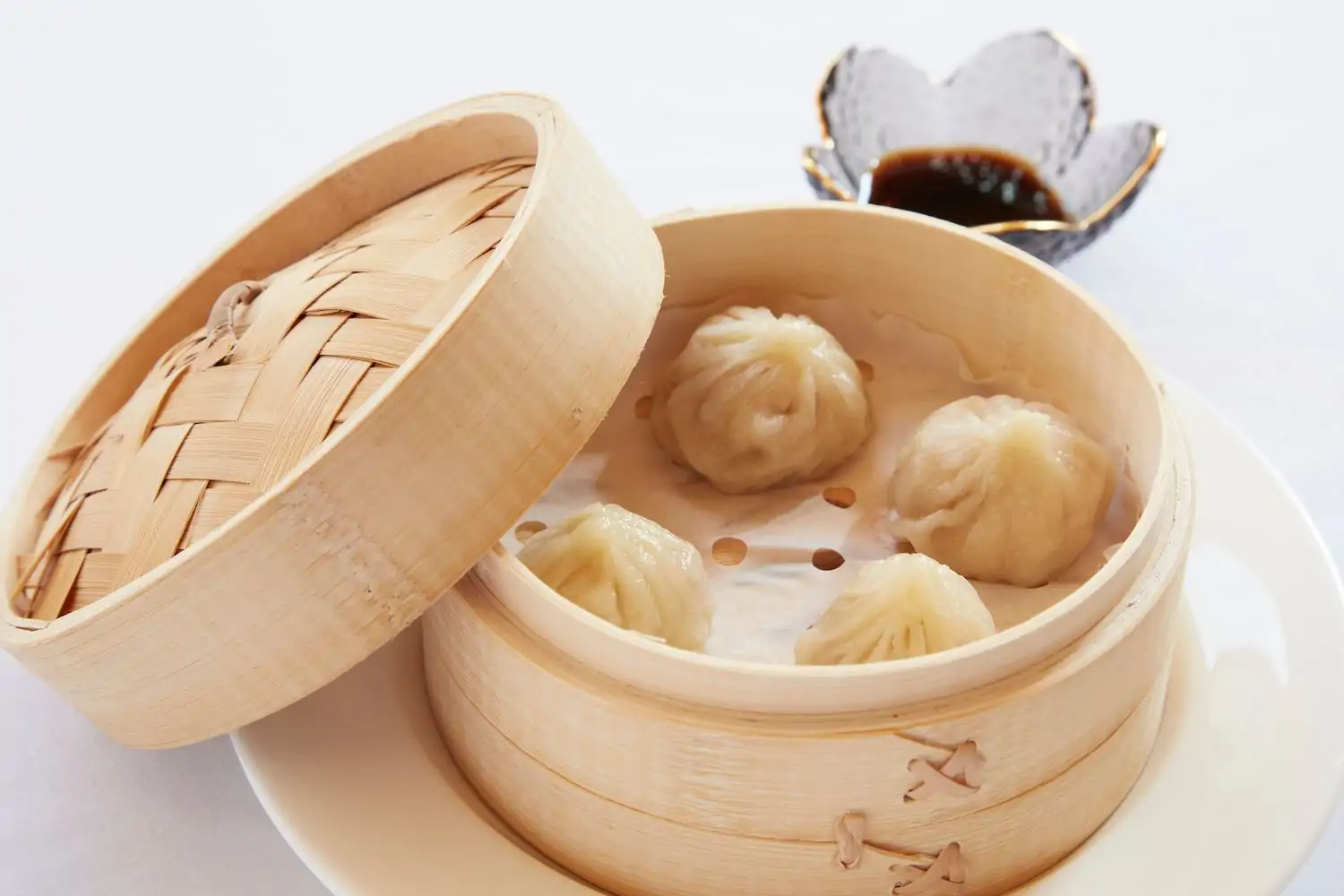 A traditional wooden container with four steamed traditional Chinese dumplings inside next to a bowl of soy sauce at Pink Moon, a glitzy Chinese restaurant in Los Angeles bookable on OpenTable.