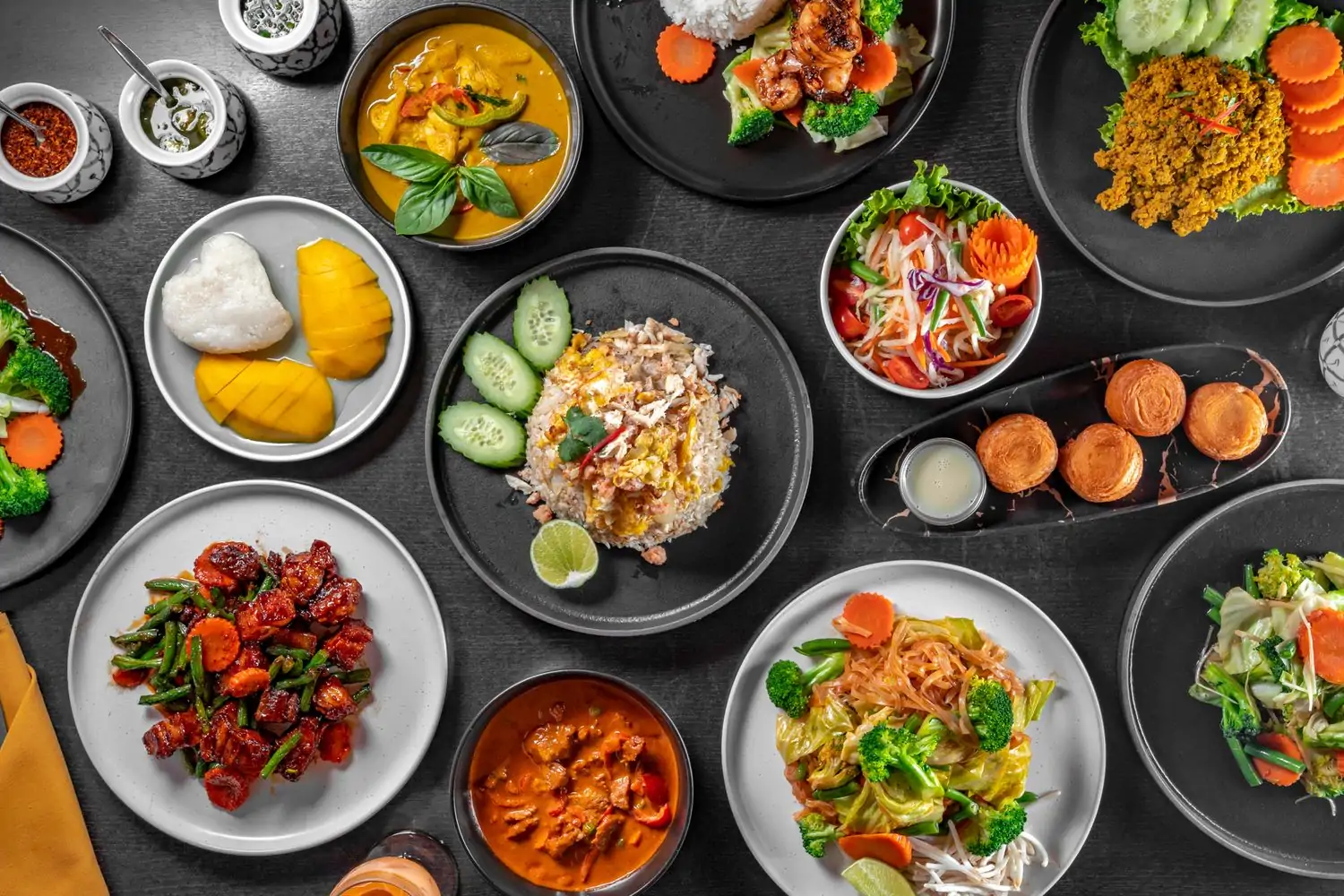 A top-down view of a black table spread with various Thai dishes, including curries, stir-fries, noodles, rice dishes, and appetizers at Emporium Thai Los Angeles, now available to book on OpenTable.