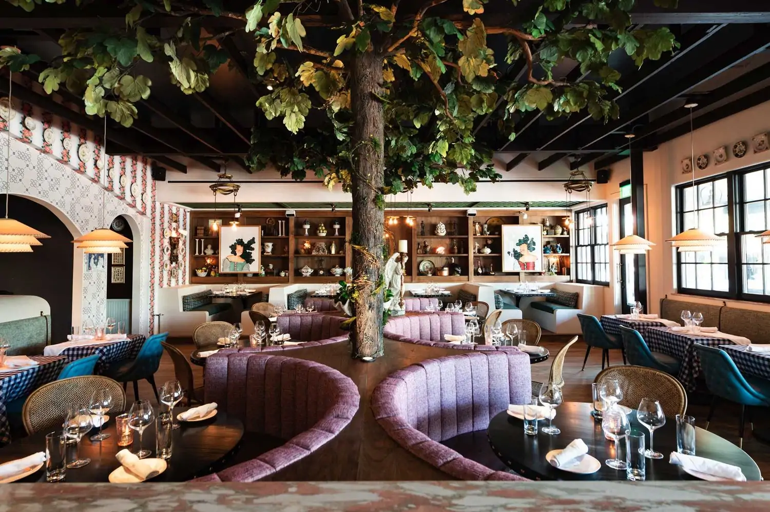 A dining room with half-circle booths, a fake tree reaching the ceiling, antiques lining the walls, and wallpaper with mosaic patterns at Sister, one of the best cozy restaurants in Dallas, on OpenTable.