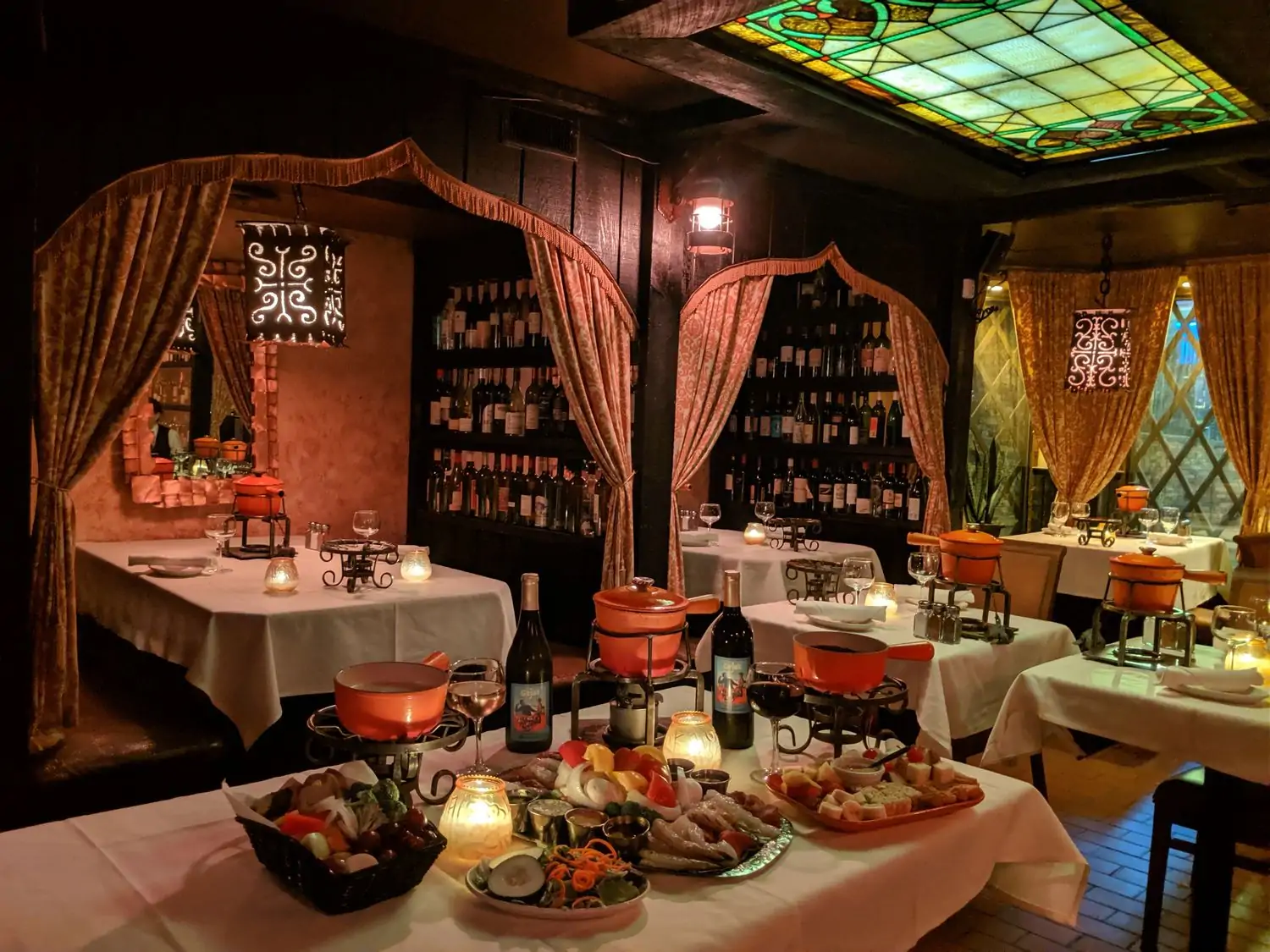 A dimly lit interior with a table setting containing bottles of wine, candles, fondue sets, and platters of veggies, meats, and cheeses with velvet curtains separating alcove tables in the background at Geja’s Cafe in Chicago found on OpenTable.