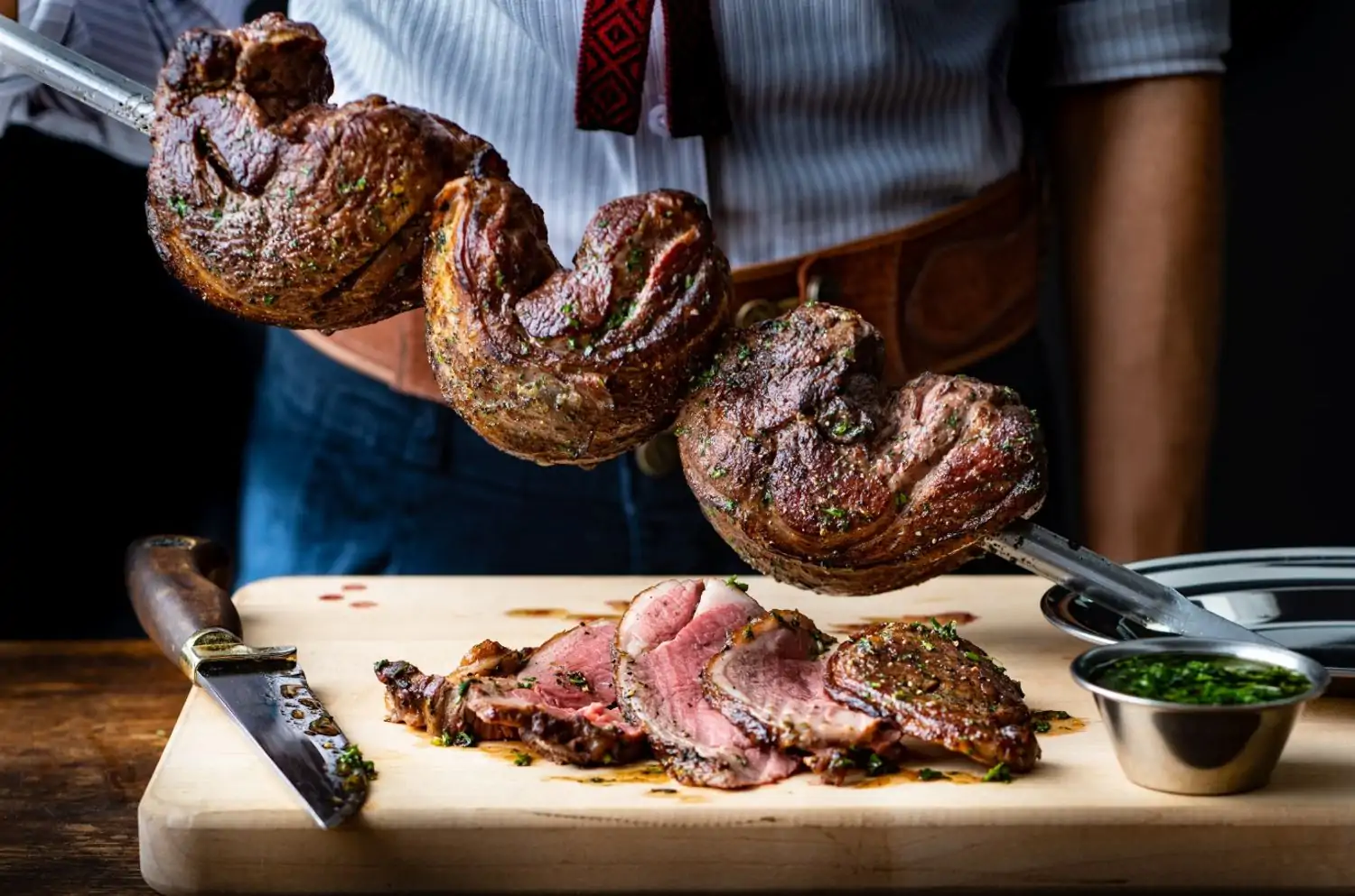 A person in a traditional Brazilian outfit carves a large, juicy piece of meat from a skewer. A plate of sliced meat and a small bowl of chimichurri sauce are also on a wooden cutting board at Fogo de Chao, one of the best Los Angeles restaurants near LAX you can reserve on OpenTable.