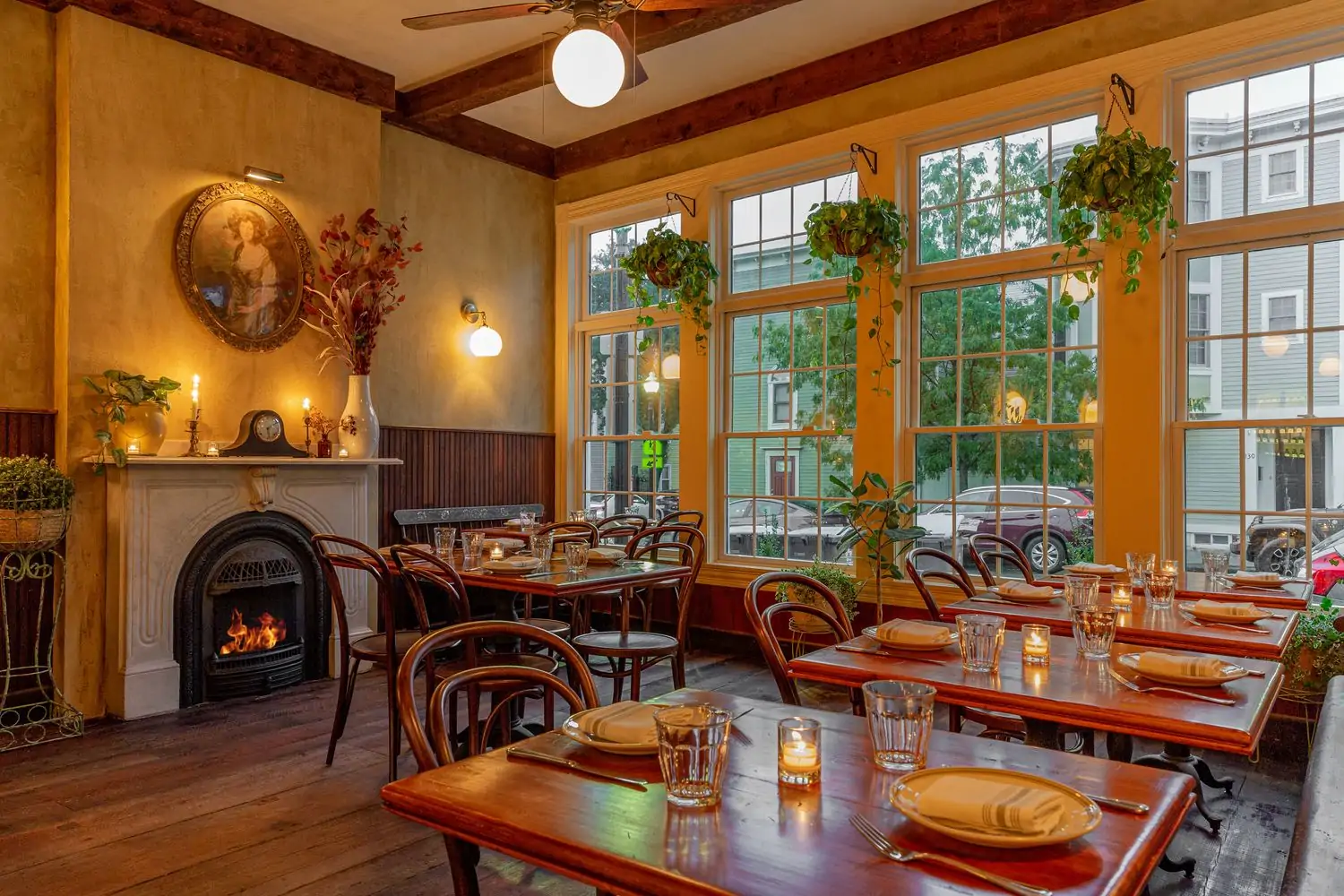 A warmly lit dining room with wooden floors and tables, a fire in the marble fireplace, a Victorian portrait on the wall, and planters hanging above the large windows at Waverly Kitchen and Bar, one of the best cozy restaurants in Boston on OpenTable.