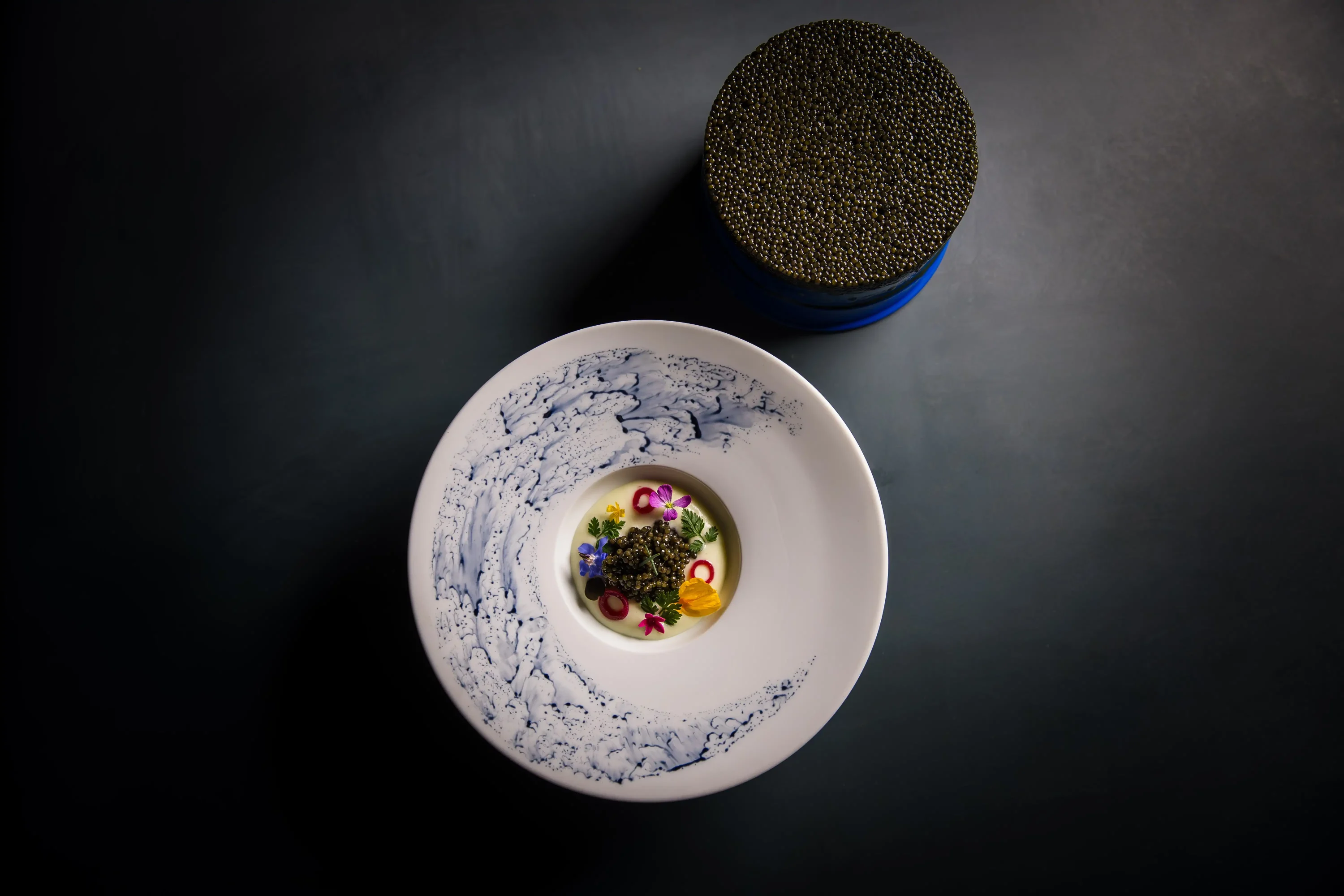 A round white plate of caviar sitting in a pale yellow broth with edible flowers and a small bowl of caviar in the background at Providence, one of the best restaurants in Los Angeles Hollywood you can reserve on OpenTable.