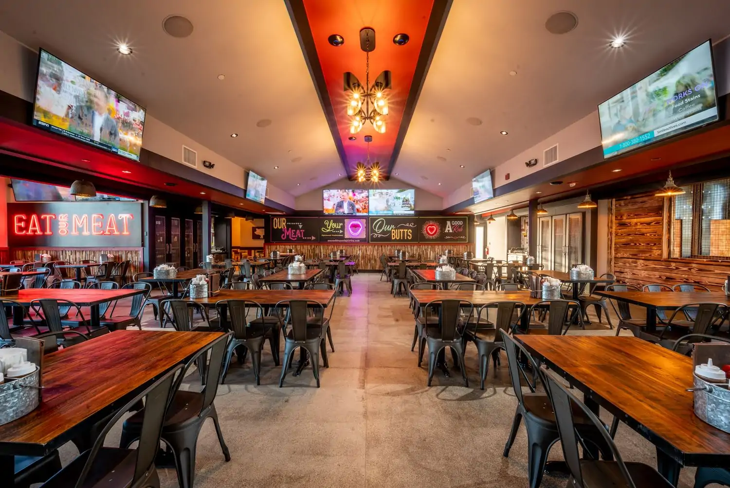 A spacious restaurant interior with wooden tables and chairs, multiple TV screens, and a red ceiling at Sauced BBQ and Spirits, one of the best Los Angeles restaurants near LAX you can book on OpenTable.