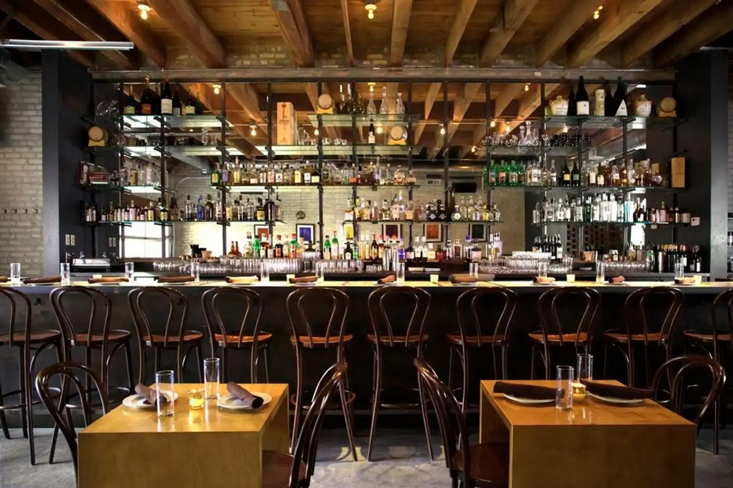 A dining room with a full bar in the background and tall, dark wood chairs in front of minimalist plate settings at Ada Street in Chicago, bookable on OpenTable.