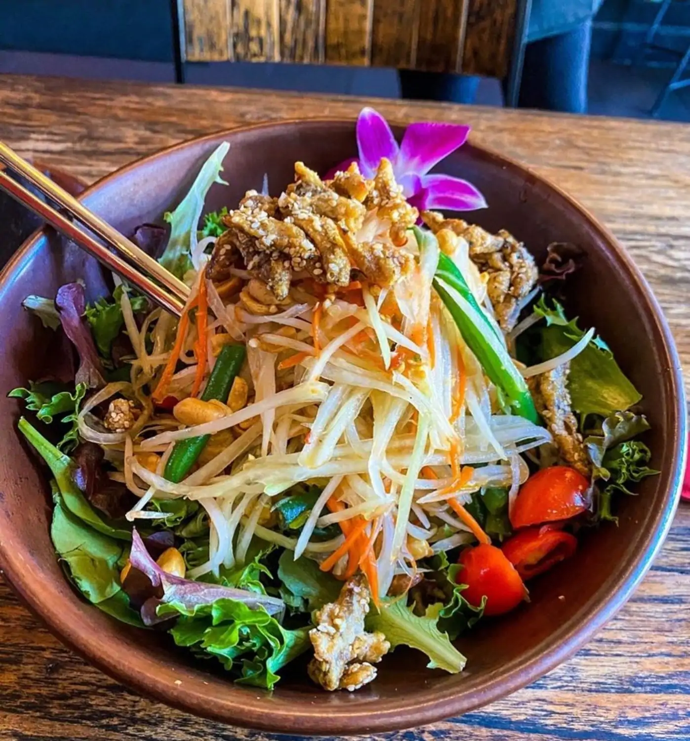 A wooden bowl of Thai papaya salad with shredded green papaya, carrots, tomatoes, peanuts, and crispy toppings garnished with a purple orchid with wooden chopsticks inside at Thai District in Los Angeles on OpenTable.