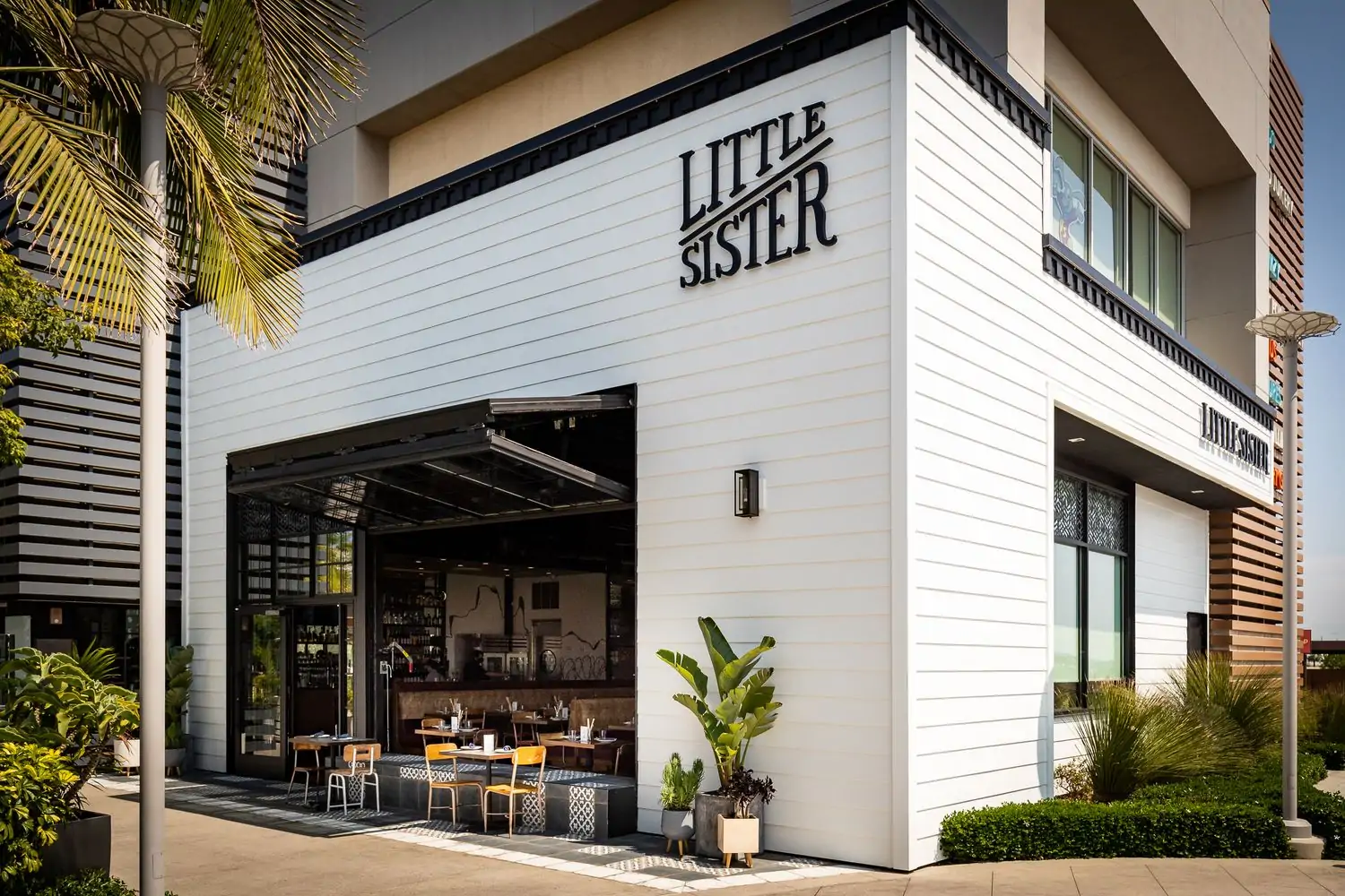 The exterior of Little Sister in El Segundo Los Angeles, a modern building with a white facade and black accents. The entrance features an oversized garage door-style window, and there are outdoor seating areas with tables and chairs, and it’s available to book on OpenTable.