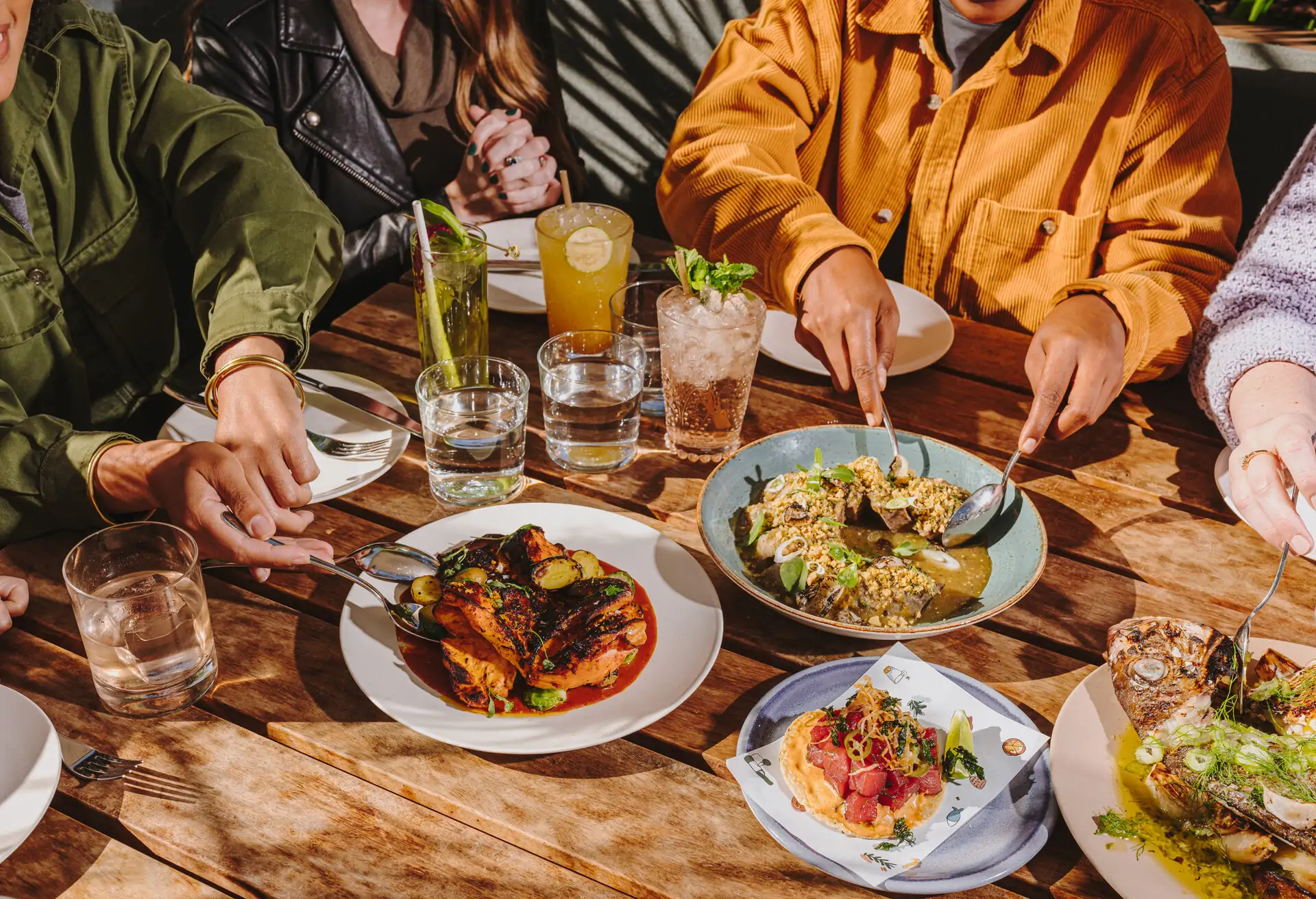A group of friends sharing colorful dishes at an outdoor table featuring plates of grilled chicken, fresh seafood, and appetizers, with cocktails and water glasses at La Cha Cha Cha, one of the top birthday restaurants in Los Angeles on OpenTable.