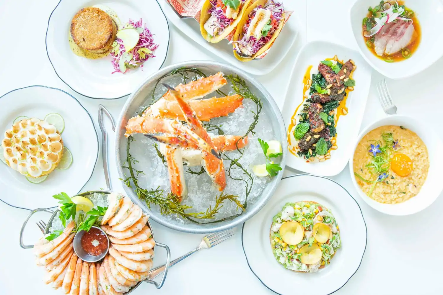 A white table displays a bowl with Alaskan king crab legs on ice surrounded by eight plates holding a variety of seafood dishes and desserts at Fishing with Dynamite, one of the best seafood restaurants in Las Angeles, on OpenTable.
