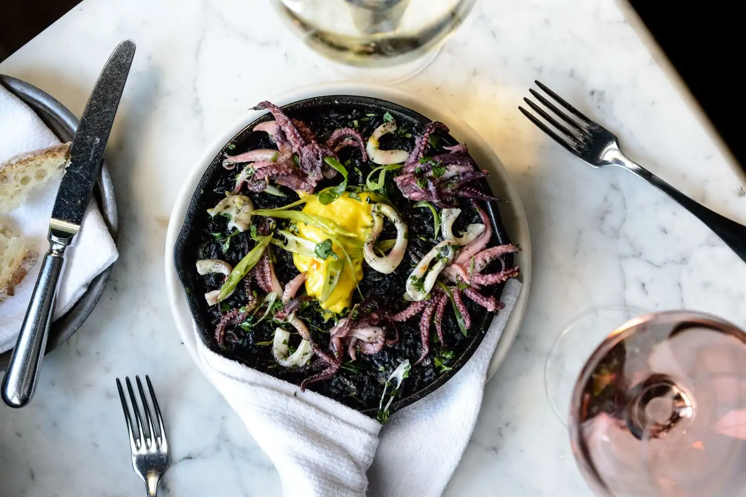 A top-down view of a black cast iron skillet filled with a dark, inky black rice dish topped with grilled octopus, green beans, and a bright yellow sauce. The dish is surrounded by a white napkin, silverware, and glasses of wine at AOC, one of the best birthday restaurants in Los Angeles you can book at OpenTable.