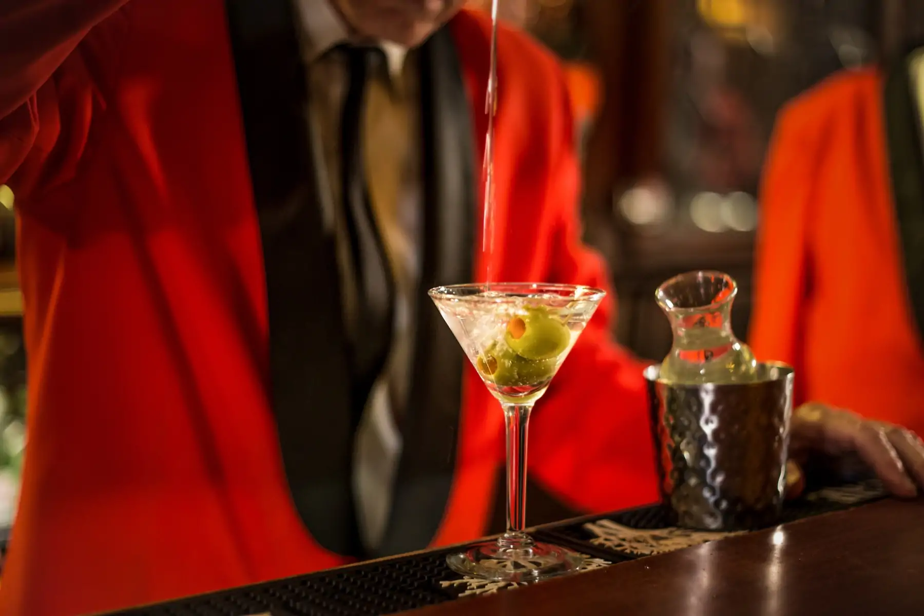 A bartender in a red jacket pouring a dirty martini into a glass with olives atop a dark wooden bar at Musso & Frank Grill in Hollywood, one of the best restaurants in Hollywood, Los Angeles, on OpenTable.