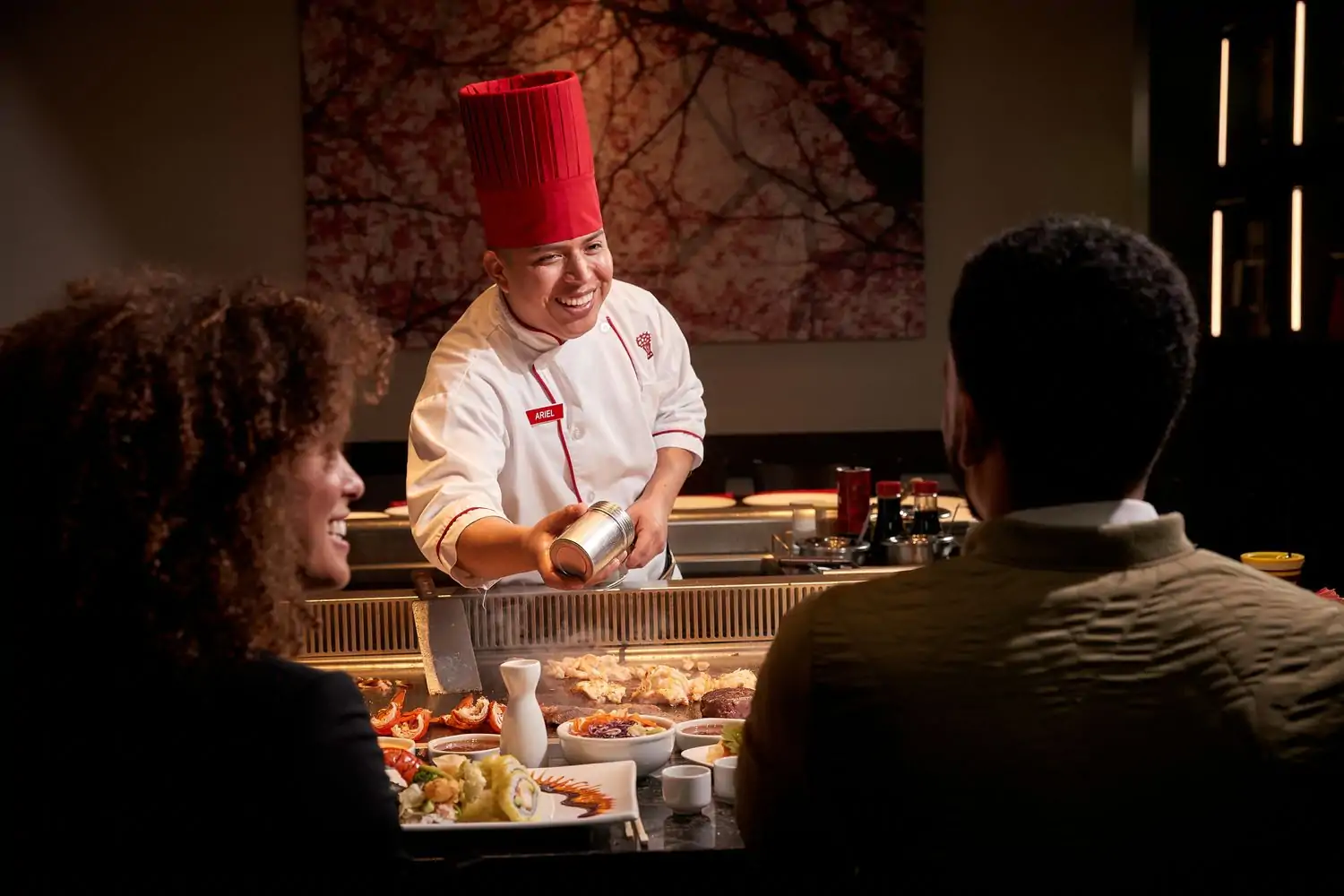 A chef in a red hat and white uniform, smiling as they engage with two guests seated at a hibachi table that’s set with a variety of colorful dishes, including sushi rolls and grilled vegetables at Benihana, one of the best birthday restaurants in Los Angeles at OpenTable.