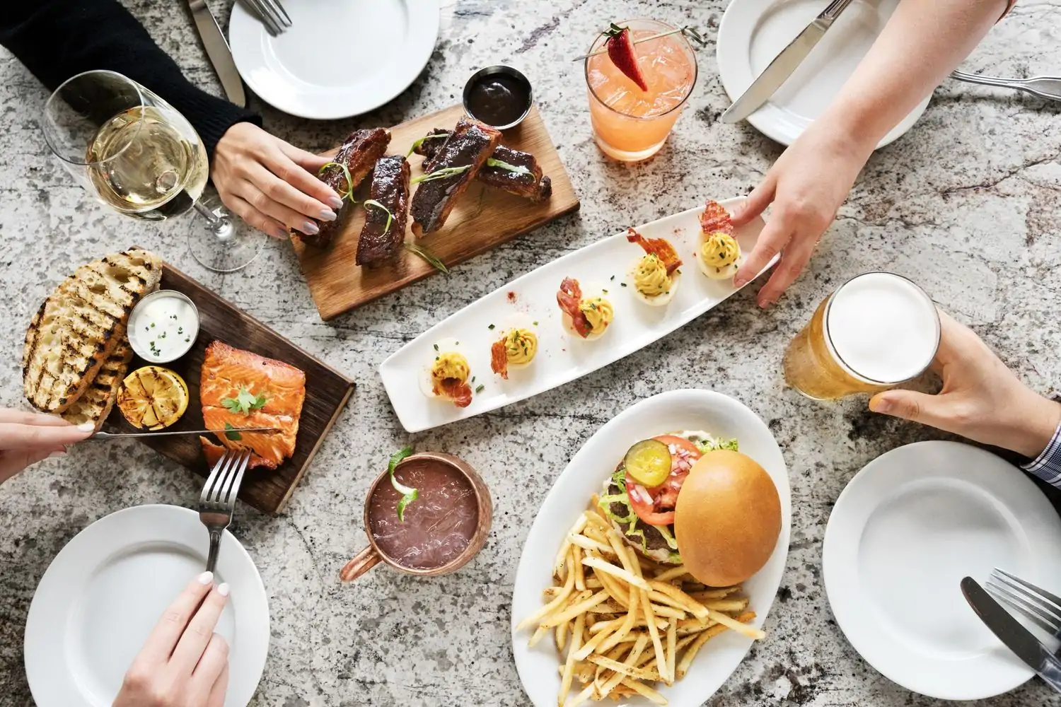 A table with a variety of food and drinks, including burgers, fries, ribs, salmon, deviled eggs, and cocktails. People's hands can be seen reaching for the food at Paul Martin’s American Grill, one of the best Los Angeles restaurants near LAX you can reserve ahead on OpenTable.