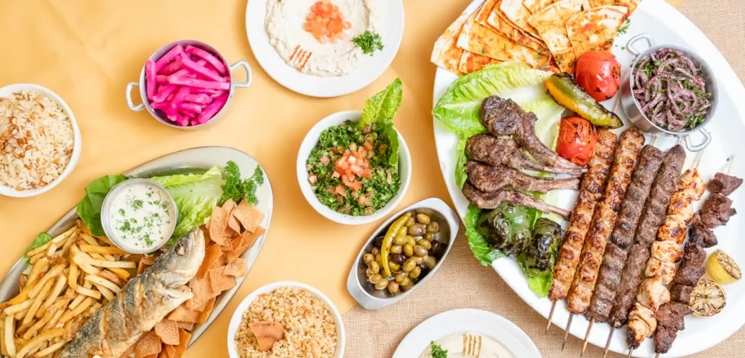A light wood table spread with a variety of Middle Eastern dishes, including grilled meats, fish, rice, salads, dips, and bread, at Carousel Restaurant, one of the best birthday restaurants Los Angeles on OpenTable.