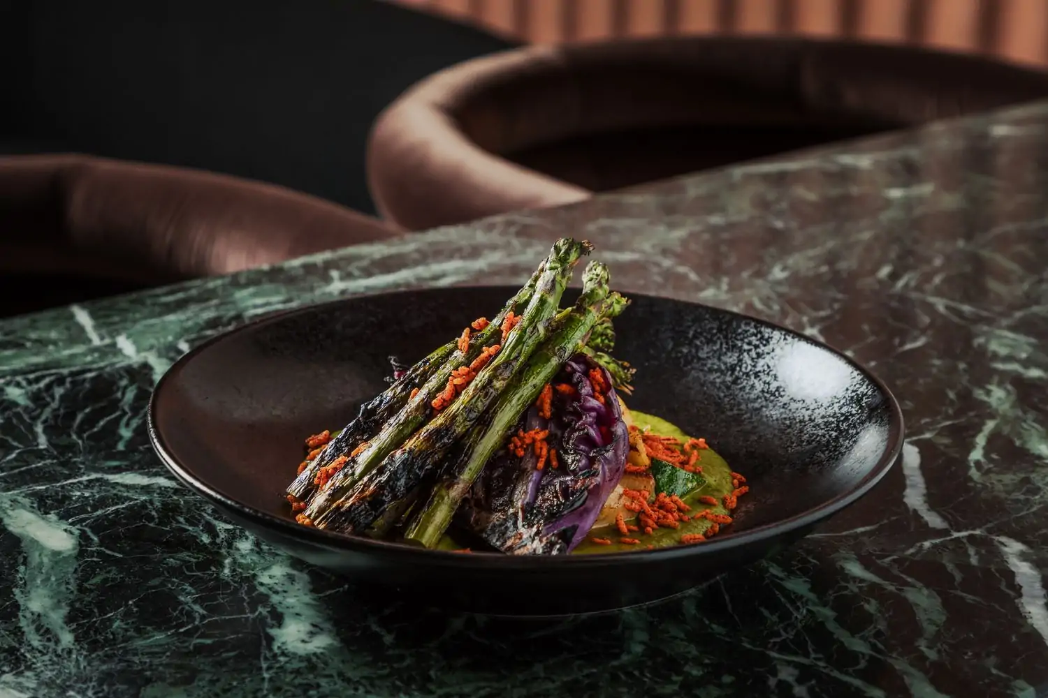 A plate of grilled asparagus and other veggies in a green sauce sitting on a marble tabletop with plush brown chairs in the background at Vela, one of the best warm and cozy restaurants in Chicago found on OpenTable.