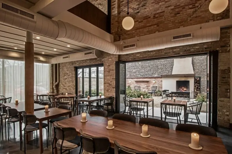 An interior dining room with red brick walls and dark wood tables with candles at the center, as well as an outdoor patio and fireplace in the background at Elske, one of the best cozy restaurants in Chicago, bookable via OpenTable.