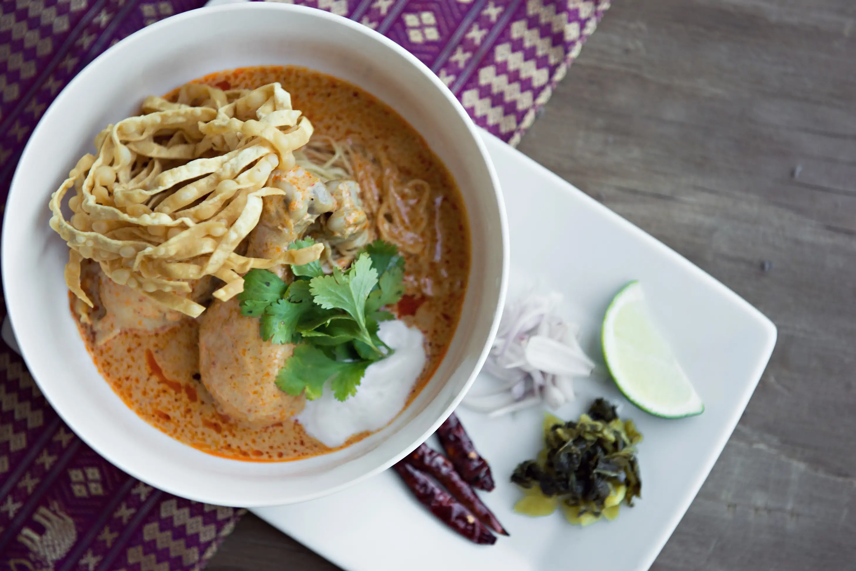 A white bowl filled with Khao Soi, a Thai noodle soup with chicken, egg noodles, cilantro, and crispy noodles with a side of lime wedge and pickled mustard greens at Ayara Thai in Los Angeles, available to book on OpenTable.