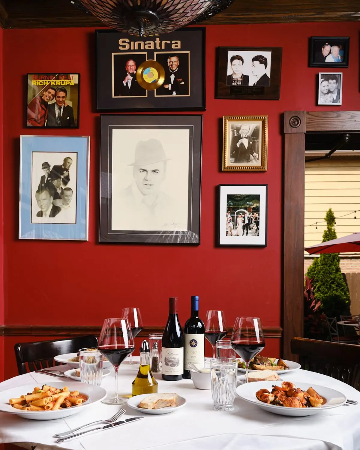 A table setting with glasses of red wine, a white tablecloth, and hearty plates of pasta in front of a deep red wall with framed photos at Mart Anthony’s in Chicago, bookable on OpenTable.