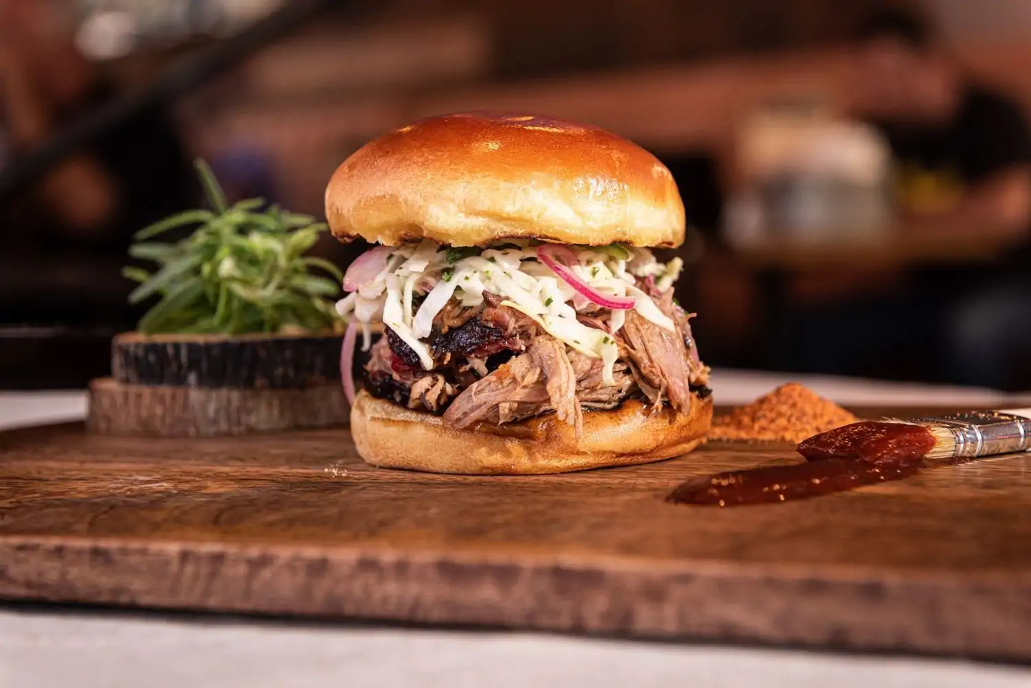 A pulled pork sandwich on a brioche bun, topped with coleslaw and drizzled with barbecue sauce on top of a wooden cutting board with a side of barbecue sauce and a sprig of rosemary at one of the best Los Angeles restaurants near LAX, Sauced BBQ and Spirits, available on OpenTable.