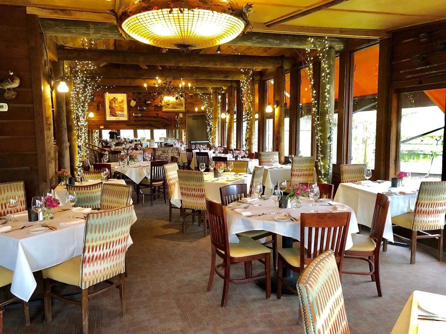 A dining room with warm wooden furnishings reminiscent of a log cabin with fairy lights around the walls and white tablecloths at Rainbow Lodge, one of the best cozy restaurants in Houston, TX.