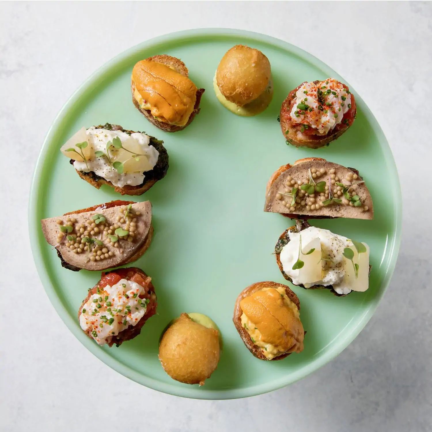 Assorted gourmet bites artfully arranged around a mint green plate, featuring a variety of toppings, textures, and flavors, including seafood, pâté, and creamy garnishes at Republique, one of the best restaurants in Hollywood Los Angeles on OpenTable.