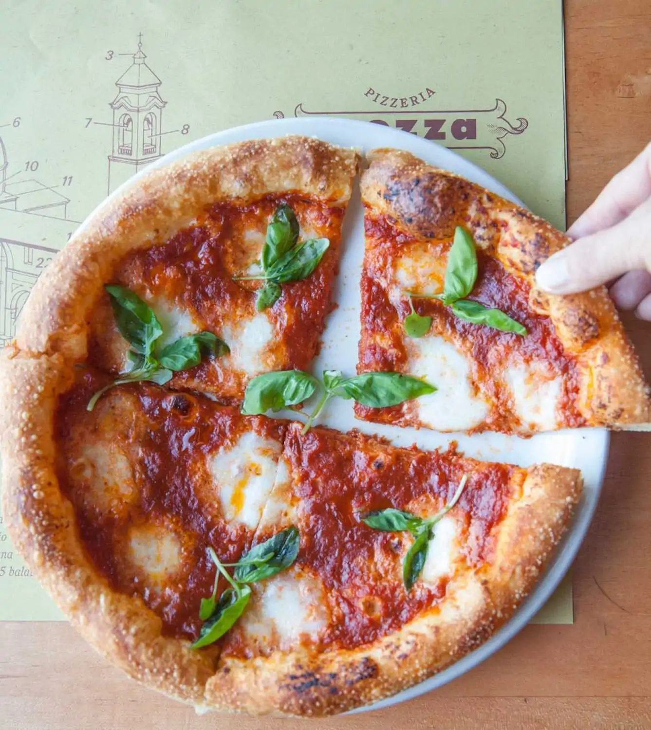 A round pizza with a thin crust topped with tomato sauce, mozzarella cheese, and fresh basil leaves. One slice is being lifted from the plate at Pizzeria Mozza in Hollywood, Los Angeles, and is available to reserve ahead on OpenTable.