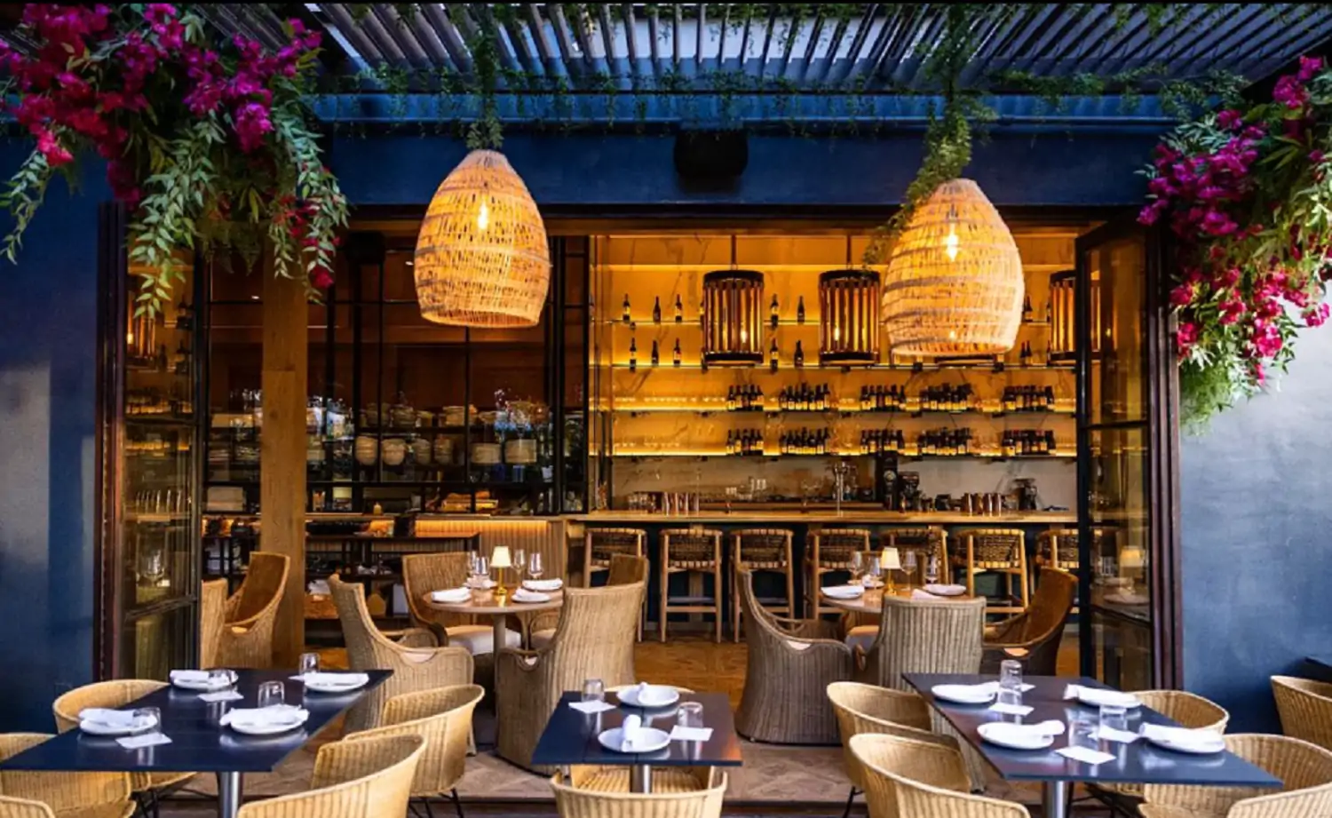 View of the covered patio dining space with a pink bougainvillea, a wooden bar, and tables surrounded by straw chairs and open glass doors of Paloma Venice, one of the best restaurants in Venice Beach, available for booking at OpenTable.