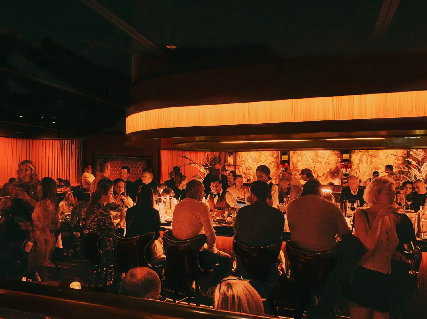 A dimly lit interior with red lighting and a crowd of people seated around the bar in high cushioned chairs at Drake’s Hollywood, one of the best cozy restaurants in Houston bookable on OpenTable.
