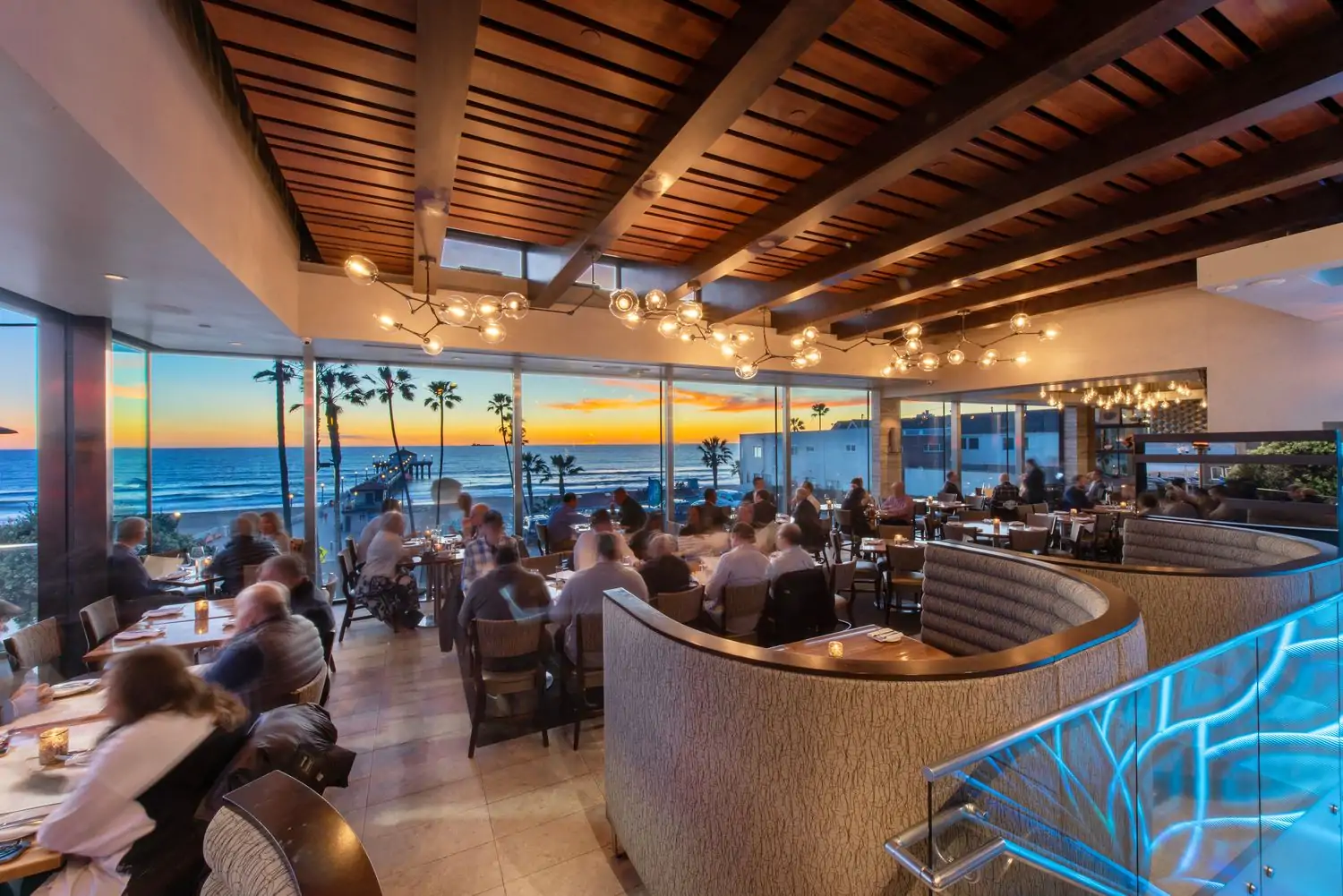 A modern dining room with warm wood accents, contemporary chandeliers, and expansive ocean views through the floor-to-ceiling windows at The Strand House, one of the best ocean-view restaurants in Los Angeles on OpenTable.