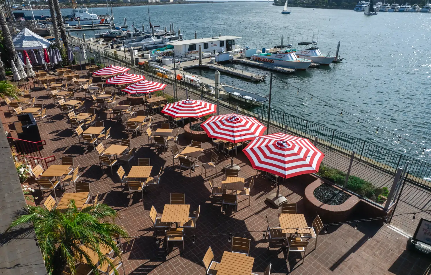 A spacious outdoor patio with tables and chairs, red and white striped umbrellas, and a view of a marina with boats and the ocean at Whiskey Red’s, one of the best beachside Los Angeles available to book on OpenTable.