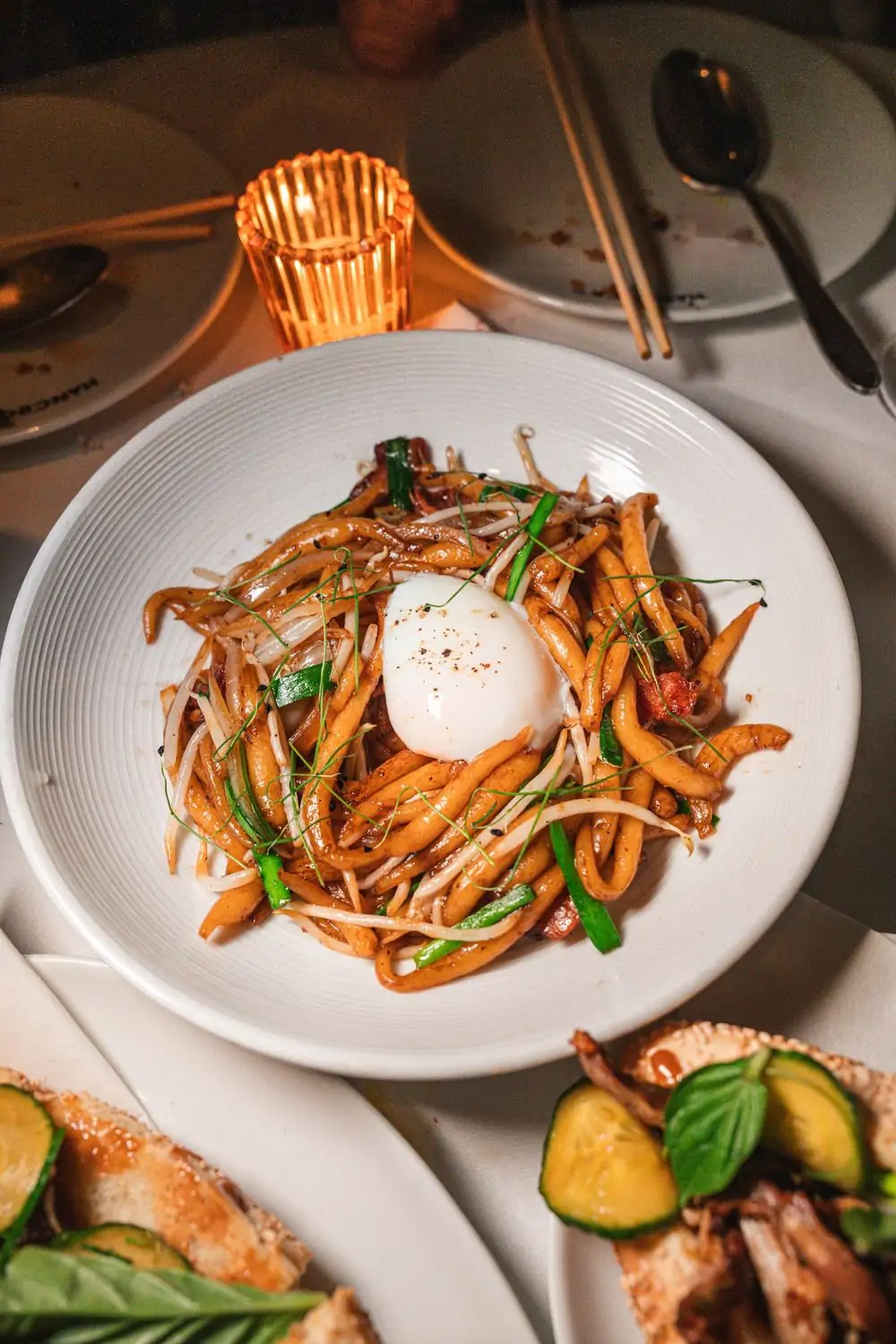 Rice noodles topped with crispy bacon, bean sprouts, garlic chives, and a soft egg rest in a white bowl next to a votive candle at Cha Cha Tang, one of the best Chinese restaurants in NYC on OpenTable.