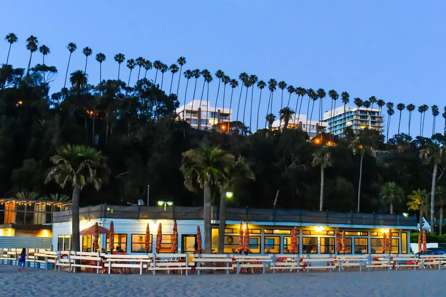 A scenic coastal view at dusk, featuring a beachfront restaurant with outdoor seating, palm trees lining the shore, and a backdrop of hills with apartment buildings at Back on the Beach, one of the best beachside Los Angeles restaurants available to book on OpenTable.