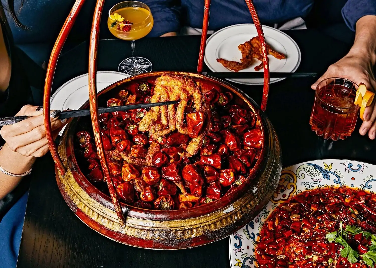 A hand holds chopsticks gripping soft shell crab in a large red bowl of dried chili peppers, surrounded by cocktails and a bowl of more chilies at Hutong, a NYC restaurant on OpenTable.