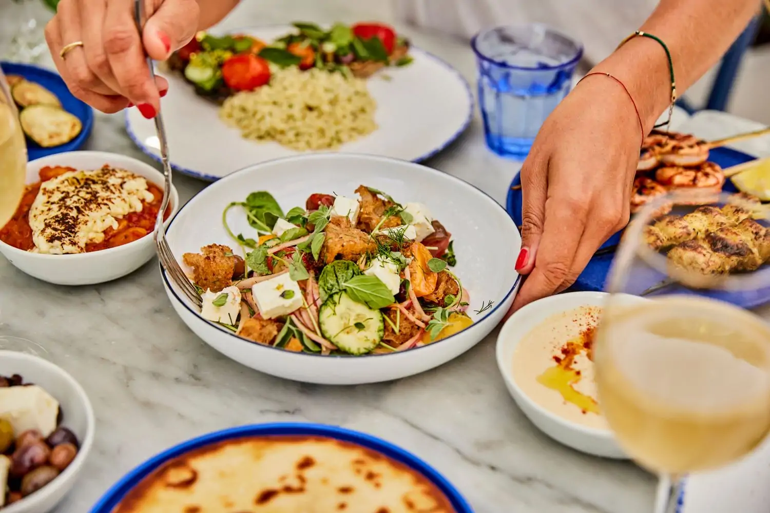 A table filled with a variety of Greek dishes, including a salad with croutons and feta cheese, a bowl of hummus, and a plate of grilled shrimp skewers with hands reaching in at Greekman’s, one of the best Silver Lake Los Angeles restaurants available on OpenTable.