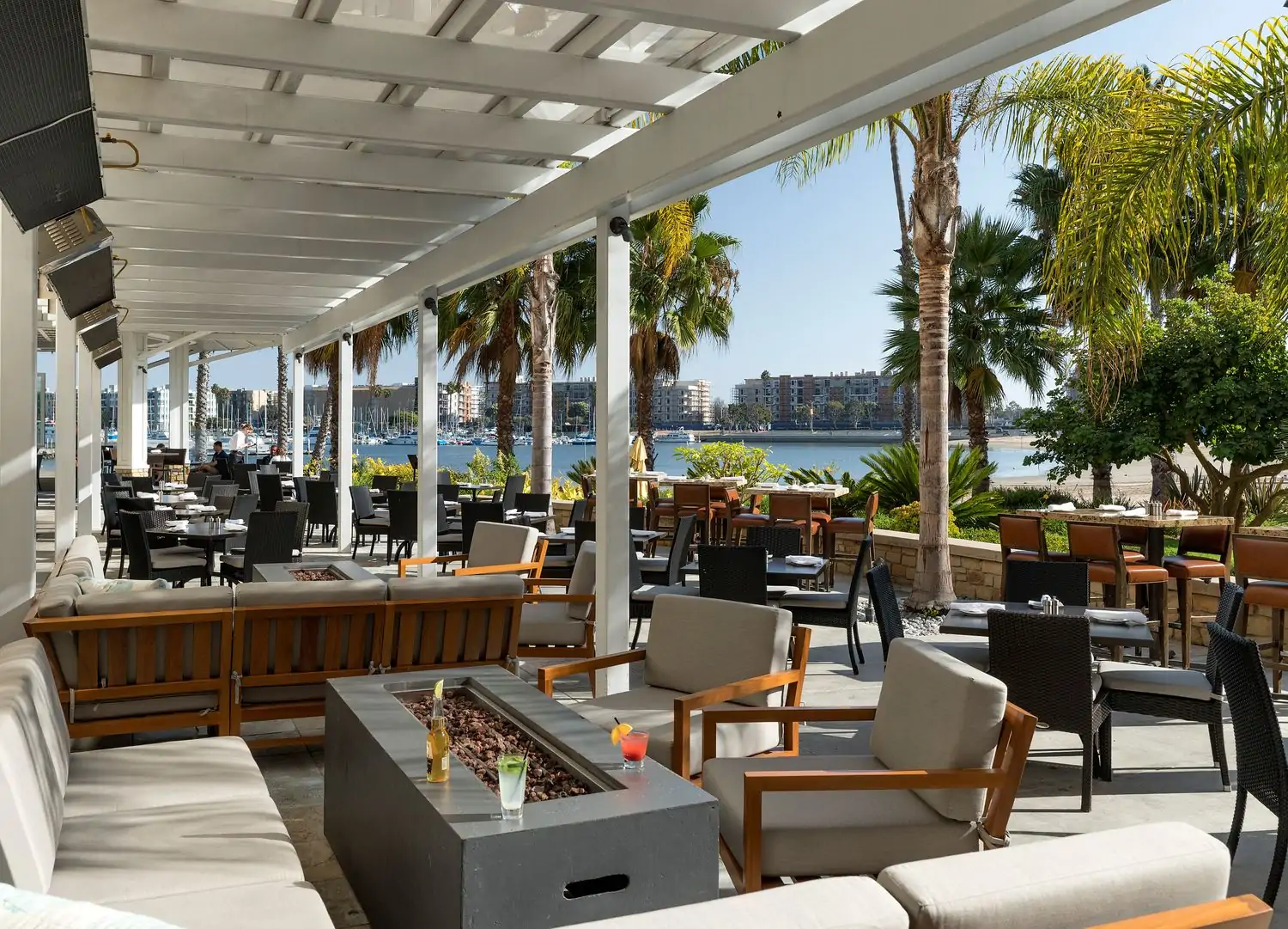 An outdoor patio with a white pergola, wooden tables and chairs, a fire pit, and a view of a marina with boats surrounded by palm stress at Beachside Restaurant and Bar in Marina del Rey, which you can book on OpenTable.
